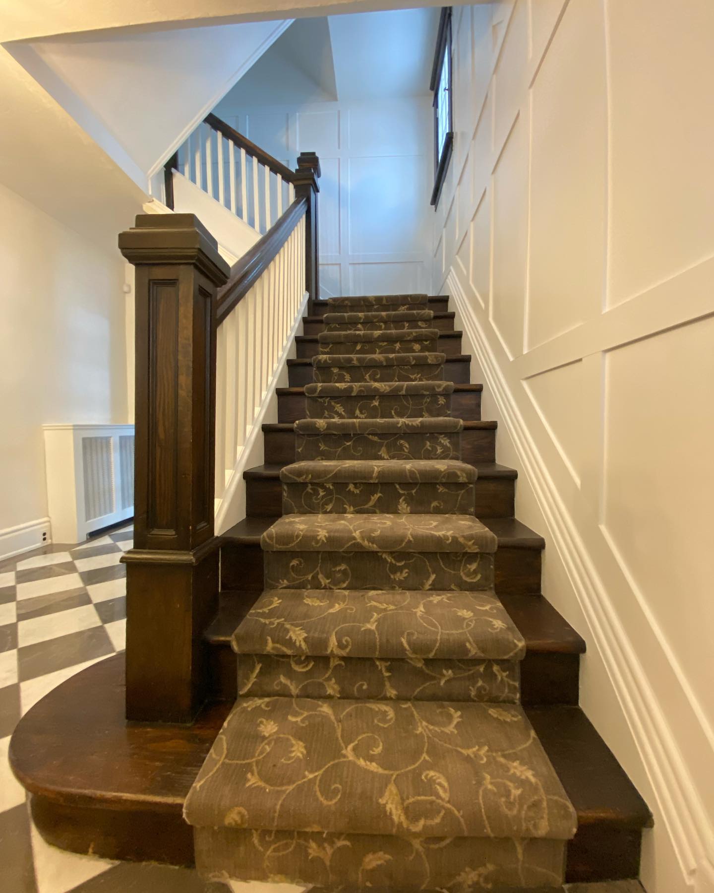 Panelled stairwell wall painted white above a dark wood newel post and carpeted stairs